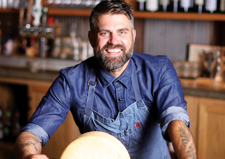 Chef Luke Zahm wears a blue striped apron and blue collared shirt, smiling behind a wheel of cheese