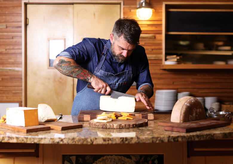 Chef Luke Zahm wears a blue striped apron and blue collared shirt. He is cutting into a wheel of cheese on a wooden table.