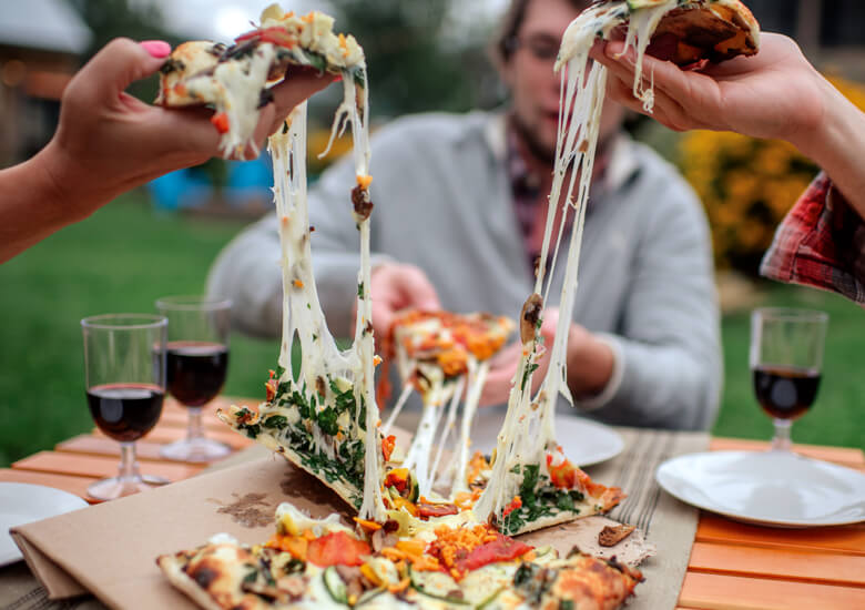 Close up of hands pulling apart cheesy pizza. A table with a pizza and drinks is seen in the background